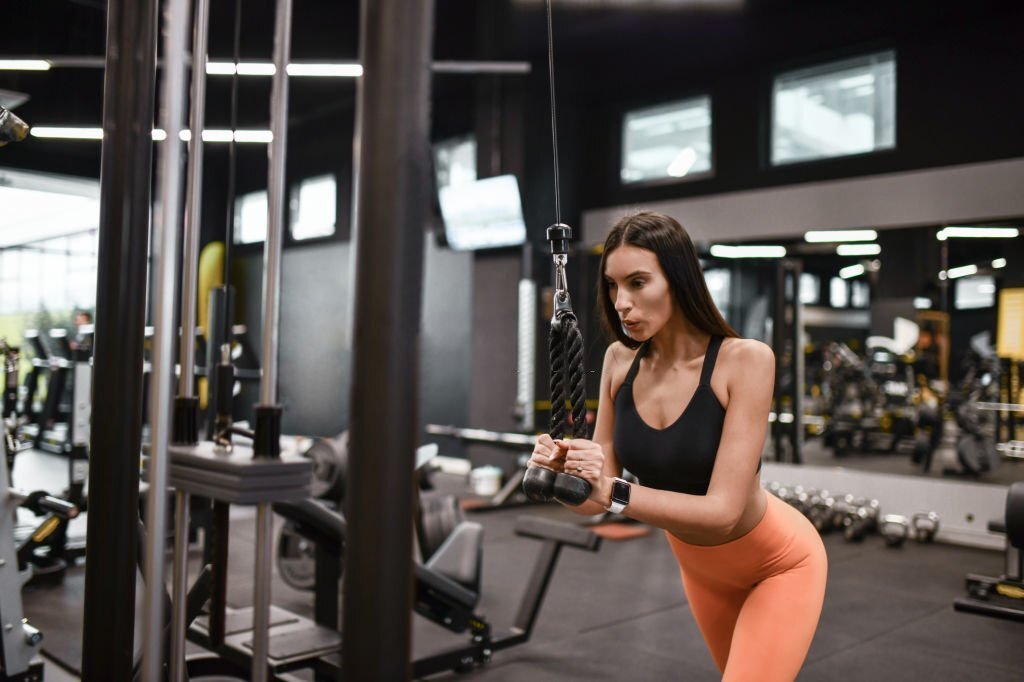 Atletė sporto salėje atlieka tricepso pratimą su jėgos treniruokliu